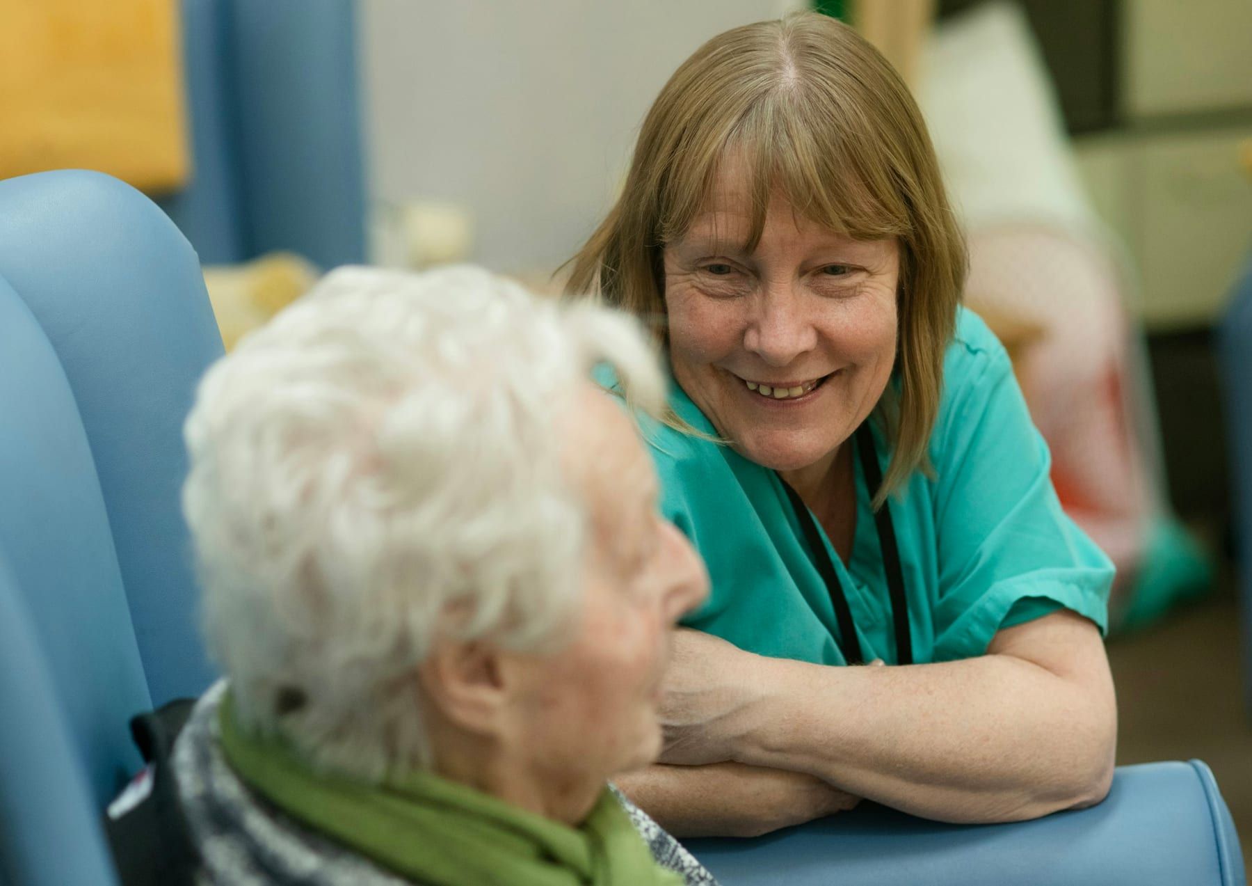A smiling caregiver in scrubs talks with an older woman