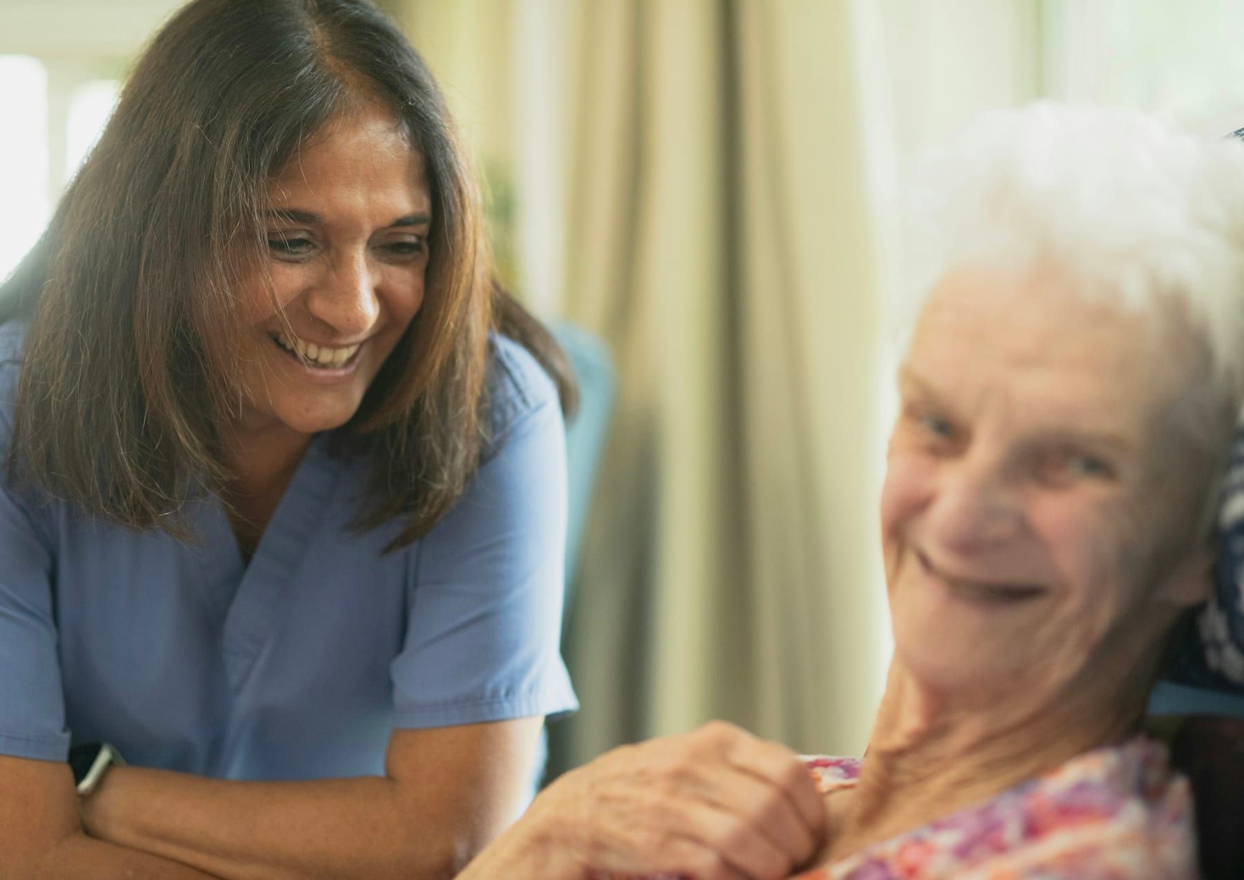A smiling caregiver leans in toward a laughing older woman