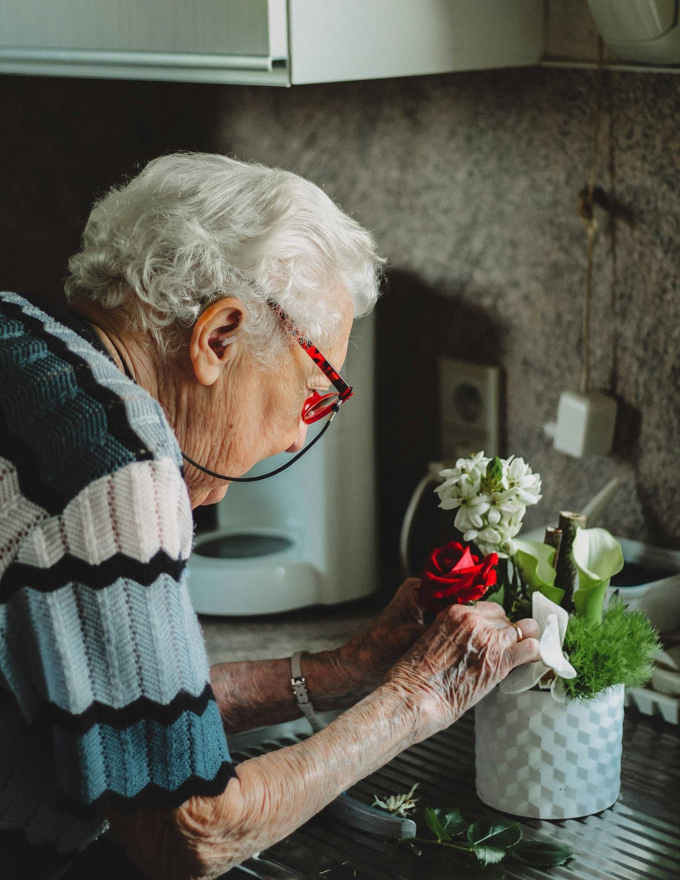 An older woman arranges flowers in a pot at her kitchen sink