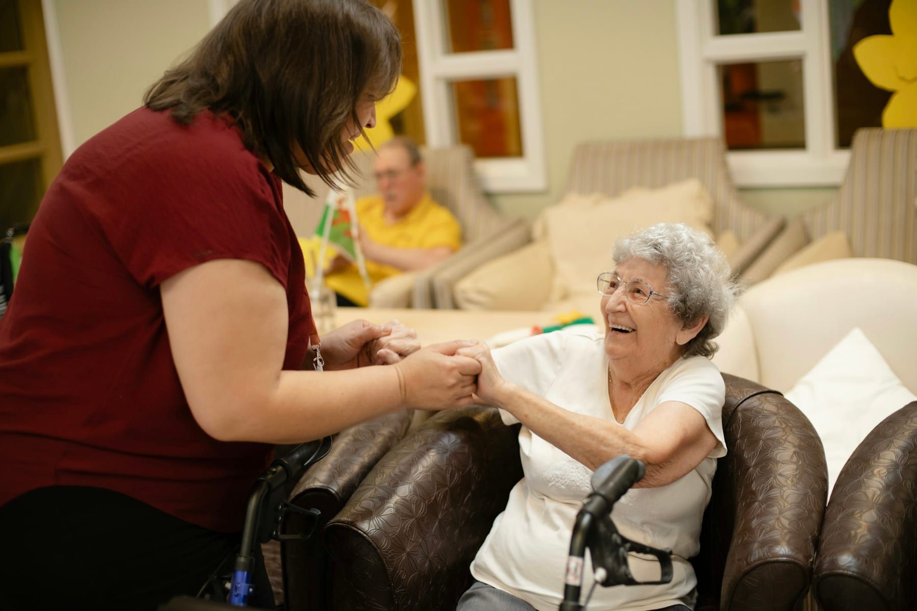 A caregiver holds hands with a smiling older woman seated beside her walker