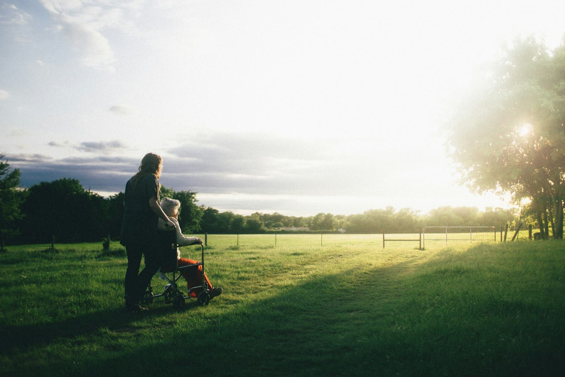 A woman walks alongside an older woman in a wheelchair across a sunny field